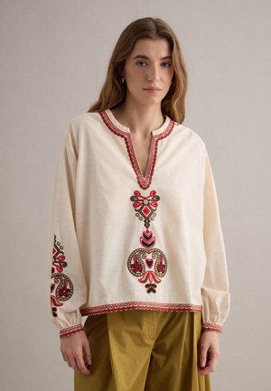 Vrouw draagt een crèmekleurige blouse met lange mouwen met rode en bruine geborduurde patronen op de borst, mouwen en zoom, gecombineerd met olijfgroene broek.