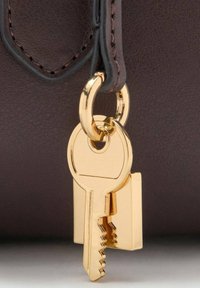 Cheie aurie atașată unui inel pentru chei, atârnând de o geantă din piele maro închis. Cheia are un design neted și plat, cu margini zimțate.