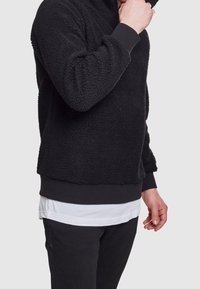 Czarny polarowy sweter z teksturowaną powierzchnią, ściągaczami na rękawach, zaokrąglonym dołem i kapturem. Nosi się go na białej, warstwowej koszuli.