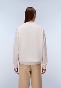 Φούτερ σε εκρού χρώμα με άνετη εφαρμογή, στρογγυλή λαιμόκοψη και ριμπ μανσέτες, σε συνδυασμό με ανοιχτό μπεζ παντελόνια, ορατά από πίσω.