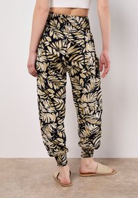 Jogging en noir et motifs dorés, avec une taille froncée et des poignets, en tissu léger et coupe décontractée. Sandales visibles.