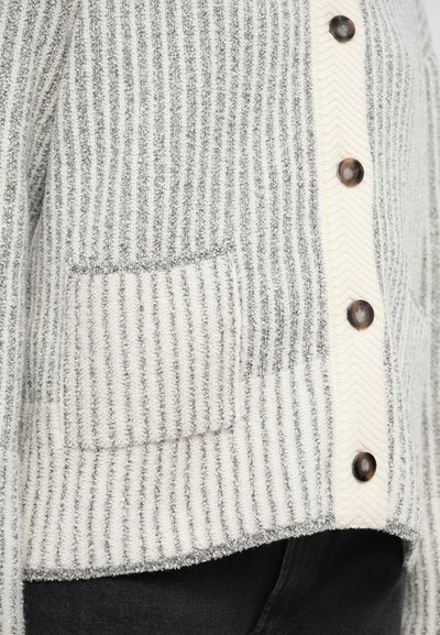 Γκρι και λευκή ριγέ ζακέτα με υφή ψαροκόκκαλο, καφέ κουμπιά και μία τσέπη στη αριστερή πλευρά.