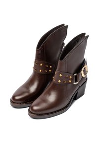 Botines de cuero marrón con punteras puntiagudas, que cuentan con un cinturón decorativo con tachuelas doradas y una cremallera lateral. Tacones de bloque y acabado texturizado.