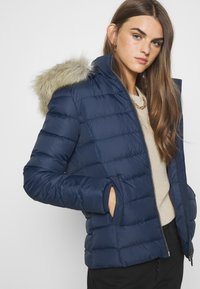 Chaqueta acolchada de color azul marino con capucha de piel sintética, cremallera frontal, bolsillos laterales y puños acanalados. Textura suave y diseño acolchado.