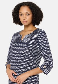 Granatowa bluzka z białym abstrakcyjnym wzorem, z dekoltem w serek i rękawami o długości do połowy przedramienia z ozdobnymi guzikami. Miękka, teksturowana tkanina.