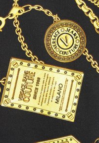 Tissu noir avec motif chaîne dorée comportant les logos et étiquettes Versace Jeans Couture, incluant du texte et des clous décoratifs.