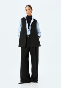 Chaleco negro sin mangas sobre una camisa azul clara, combinado con pantalones amplios negros. El conjunto incluye un cinturón y un cuello de tortuga negro texturizado.