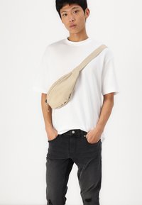 Beige stoff krossbody-veske med rund form og glidelås, bæres over en ensfarget hvit t-skjorte og kombinert med slitte svarte jeans.