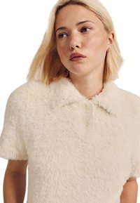 Polo en peluche beige avec une texture douce, manches courtes et patte à trois boutons. Doté d'un col classique et d'une subtile brillance.
