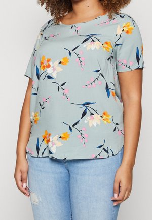 Mujer con una blusa azul claro de manga corta con estampado floral en amarillo, blanco y rosa, combinada con vaqueros azules claros rasgados.