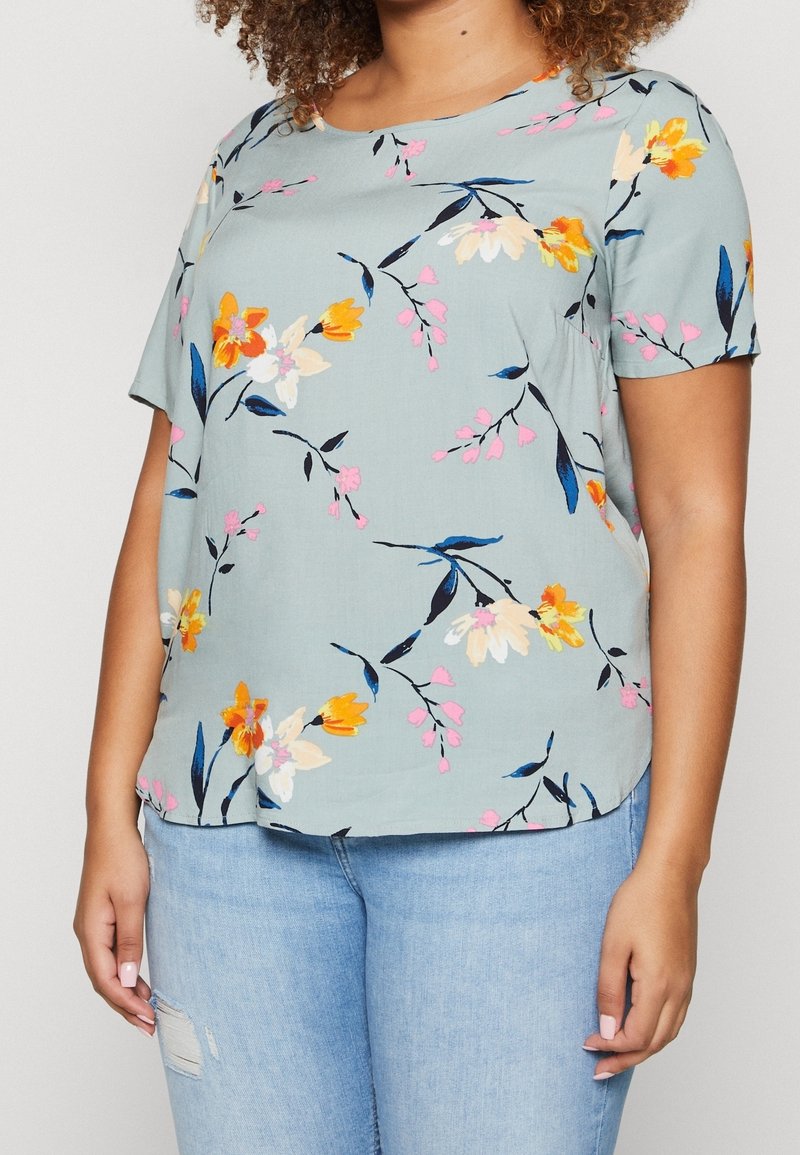 Femme portant un chemisier bleu clair à manches courtes avec un imprimé floral jaune, blanc et rose, associé à un jean bleu clair déchiré.