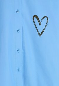 Jasnoniebieska, bawełniana koszula zapinana na guziki, z czarnym sercem na lewej klatce piersiowej, niebieskimi guzikami i gładką fakturą.