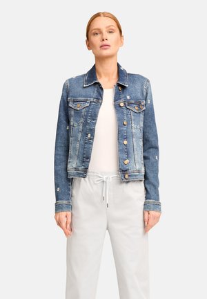 Woman wearing a blue denim jacket with small white daisy embroidery, white top, and light gray drawstring pants, standing against a white background.