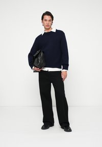 Mies, joka käyttää laivansinistä villapaitaa valkoisen paidan päällä, mustia leveälahkeisia housuja, mustia kenkiä ja pitää suurta mustaa nahkakäsilaukkua valkoista taustaa vasten.