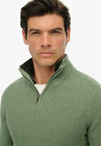 Grüner Strickpullover mit einem strukturierten Muster, halber Reißverschluss am Kragen und einem dunklen Karomuster im Krageninneren.