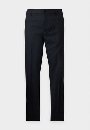 Homme portant un pull violet doux, un pantalon noir et des baskets blanches, debout avec une main dans la poche devant un fond uni.