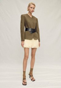 Olive green blazer with structured shoulders, black wide belt, layered cream ruffled mini skirt, sheer brown socks, and black heeled sandals.