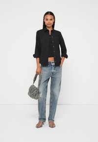 Melna pogājama krekla virsī īss melns tops, kombinēts ar gaiši zilām nodiluma džinsu biksēm un pelēku somiņu ar rakstu. Modele valkā atvērtu purngalu sandaļus.