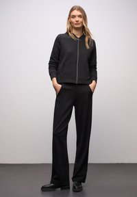 Zwarte doorgestikte jas met ritssluiting en staande kraag, gecombineerd met loszittende zwarte broek en stevige zwarte laarzen. Soepele textuur en eenvoudig ontwerp.