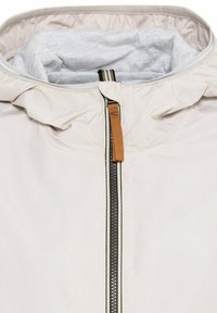 Lys beige jakke med høj krave, der har en sort lynlås og en brun tag. Indvendigt viser den en lys grå foring og stribede detaljer.