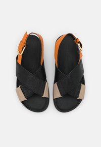 Mustat ja oranssit sandaalit, joissa on teksturoitu ristimääräysdesign, beige hihna ja kultainen solkiaksentti. Tasapohja.