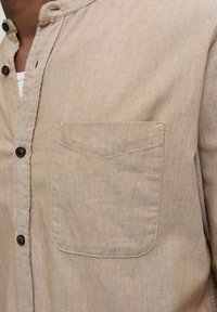 Light brown button-up shirt with a small front pocket, made from a textured fabric. Features a mandarin collar and two buttons near the neck.