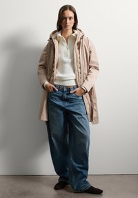 Giacca puffer beige con cappuccio, maglione bianco a coste e jeans blu a gamba larga. Scarpe nere slip-on completano l'outfit.