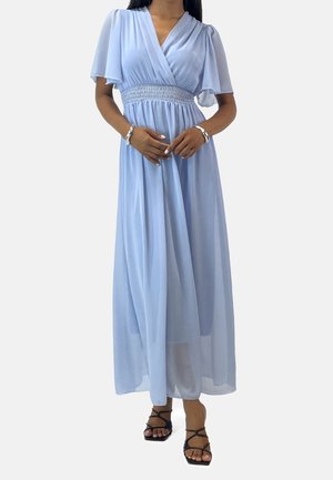 Femme portant une robe maxi fluide bleu clair avec des manches papillon, une taille froncée, et des sandales à talons noires à brides sur fond blanc.