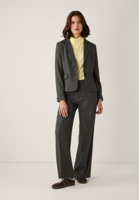 Grauer strukturierter Anzug mit einer taillierten Blazerjacke und weiten Hosen, kombiniert mit einem hellgelben Rollkragenpullover. Braunes Schuhwerk rundet den Look ab.