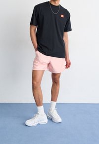 Man med svart t-shirt med röd lapp, ljusrosa shorts, vita knästrumpor, vita sneakers och silverkedja, stående på blått golv.