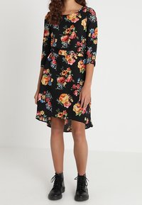 Robe fleurie à manches trois-quarts, base noire, motif de roses et de fleurs vibrantes, ourlet asymétrique, associée à des bottines noires.