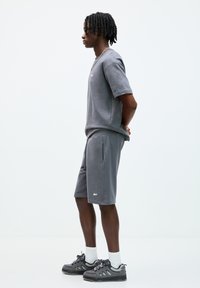 Camicetta a maniche corte grigia in cotone e pantaloni corti coordinati, con un logo minimale. Abbinata a scarpe sportive scure e calzini bianchi fino alla caviglia.
