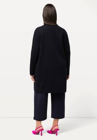 Lång svart cardigan med rak skärning, parat med marinblå capribyxor och klara rosa klackar, vilket framhäver en kontrast i färg och textur.
