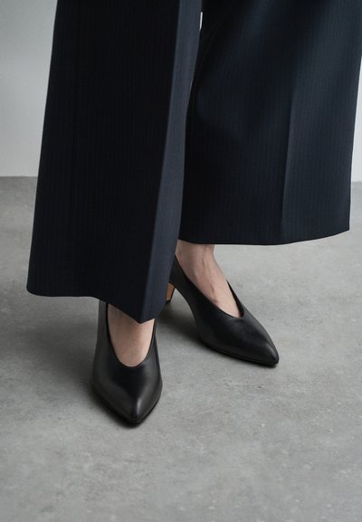 Scarpe con tacco nere in pelle con punta a cono e tacco basso quadrato, abbinate a pantaloni scuri a righe sottili su un pavimento in cemento.