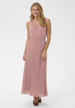 Uśmiechnięta kobieta o długich blond włosach, ubrana w bezrękawową plisowaną sukienkę maxi w kolorze matowego różu oraz brązowe plecione szpilki na tle jednolitego tła.