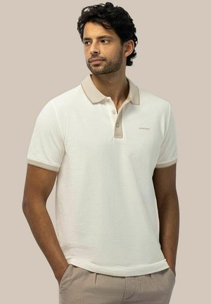Man with short curly hair wearing a white polo shirt with beige collar and sleeve edges, standing with hands in beige pants pockets.