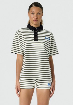 Vrouw die een wit shirt met korte mouwen en korte broek draagt met zwarte horizontale strepen en een zwarte kraag, staand tegen een effen achtergrond.