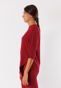 Donna con capelli castani intrecciati, che indossa un top rosso a tre quarti e pantaloni rossi abbinati, in piedi di profilo contro uno sfondo bianco.