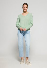 Mintgrön v-ringad tröja i mjukt stickat material, kombinerad med ljusblå jeans och tan sneakers, med en diskret logotypdetalj på bröstet.