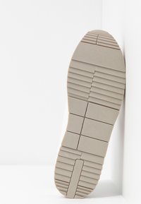 Semelle de chaussure beige clair avec des motifs de bande de roulement horizontaux et rectangulaires, positionnée verticalement sur un fond blanc en coin.