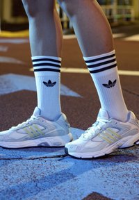 Bijele sportske tenisice s žutim akcentima i teksturiranom mrežicom, uparene s bijelim čarapama s crnim prugama i Adidas logom.