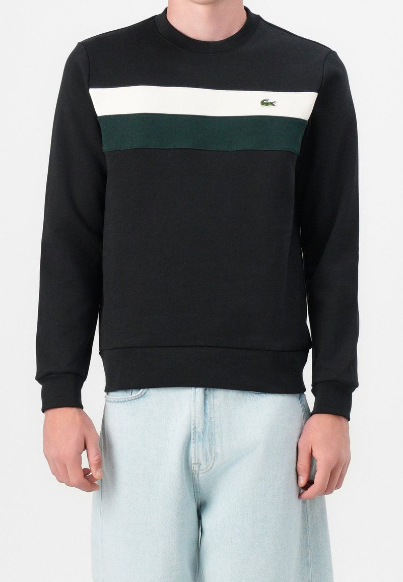 Homme portant un sweat-shirt noir avec des rayures horizontales blanches et vert foncé et un logo sur la poitrine, associé à un jean bleu clair.