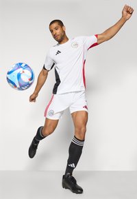 Maillot de football blanc avec des accents noirs et rouges, complété par un short blanc. Le joueur porte des chaussures noires et des chaussettes noires montantes avec des rayures blanches.