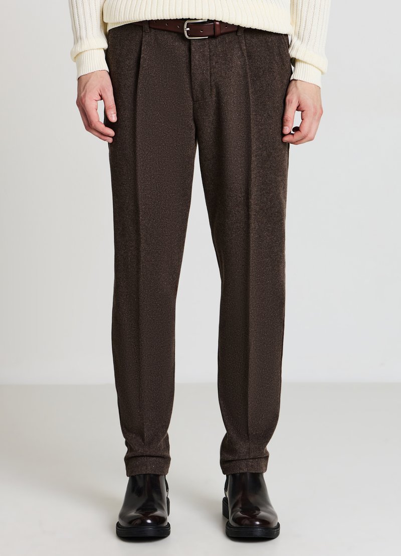 Braune, plissierte Hose aus strukturiertem Stoff. Am Bund mit einem braunen Gürtel eng anliegend, in Knöchellänge, kombiniert mit schwarzen Schuhen.