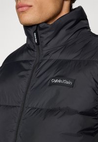 Μαύρο μπουφάν πούφ με ψηλό γιακά, που διαθέτει φερμουάρ μπροστά και ορθογώνια ετικέτα λογότυπου Calvin Klein στο στήθος.