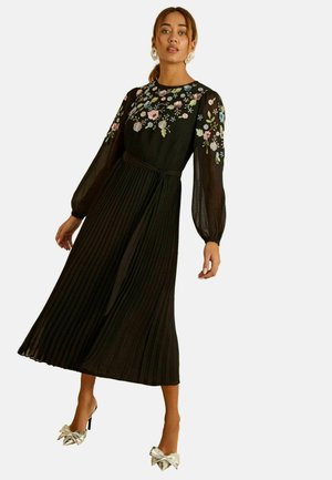 Fekete pliszírozott ruha hosszú, áttetsző ujjakkal és virágos hímzéssel. Tartozik hozzá egy hozzáillő deréköv, és ezüst masnimosdattal díszített magassarkú cipővel párosítva.