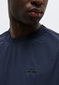 Detailný záber na osobu oblečenú v tmavomodrej košeli s malým čiernym logom na hrudi, na fotografii je viditeľná časť krku a pier.