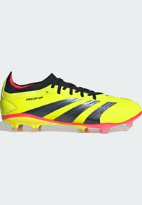 Chaussure de foot jaune avec des accents noirs et rouges, comportant un revêtement texturé, un système de laçage et des crampons translucides pour une meilleure adhérence.