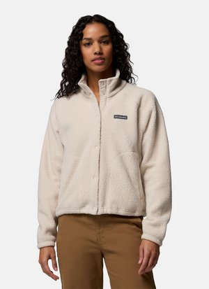 Kvinde iført en cremefarvet fleecejakke med opretstående krave og brune bukser, stående mod en ensfarvet baggrund.