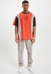 Overdimensjonert t-skjorte med oransje, grå og svarte paneler; har "ECKŌ"-logo. Grå bukser med sidesømmer og hvite sneakers.